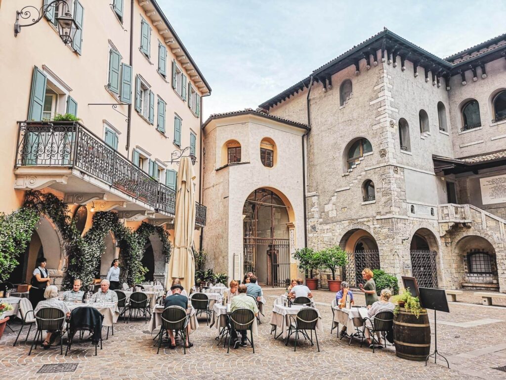 Riva del Garda am nördlichen Gardasee in Italien: Das Bild zeigt die Piazza San Rocco in der Altstadt von Riva del Garda mit Außentischen eines Restaurants, umgeben von historischen Gebäuden aus Stein