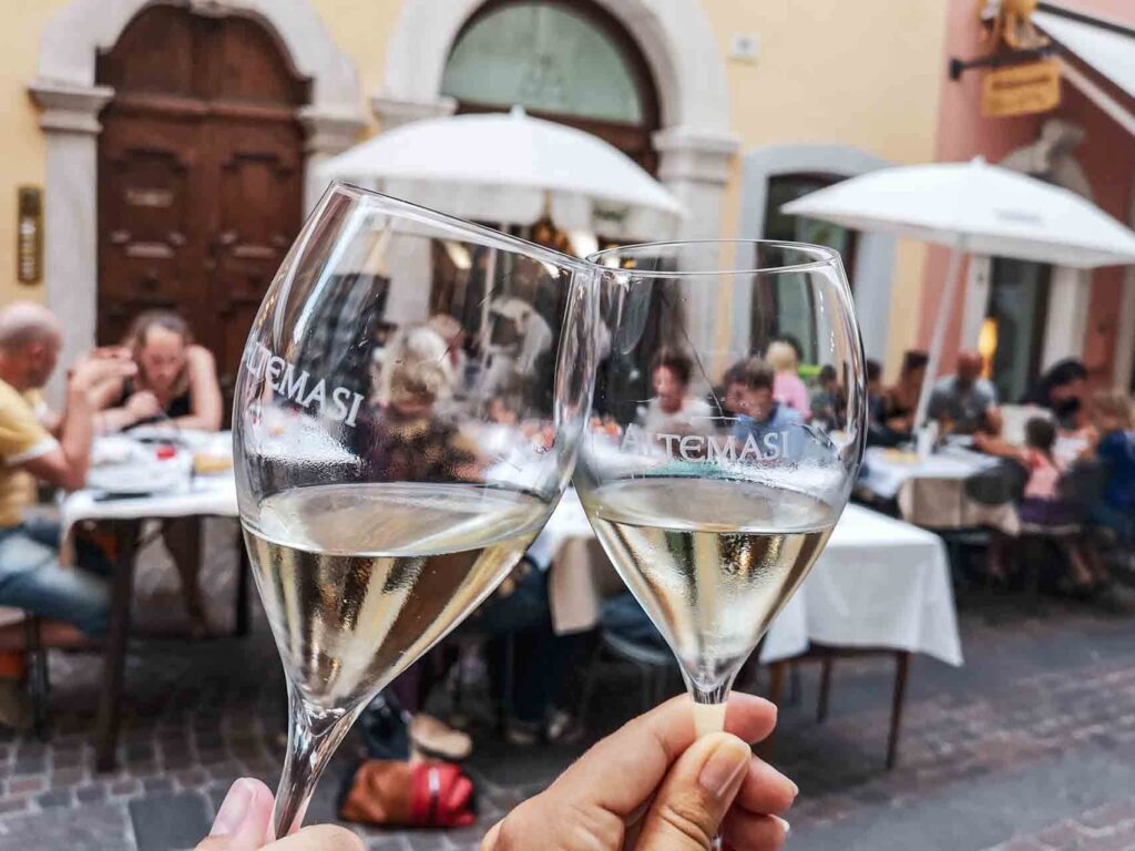 Restauranttipp für Riva del Garda am nördlichen Gardasee in Italien: Aperitif im Ristorante Leon d'Oro. Auf dem Bild sind zwei Gläser mit Prosecco beim Anstoßen zu sehen, im Hintergrund die Tische des Restaurants im Freien in einer engen Gasse in der Altstadt von Riva del Garda