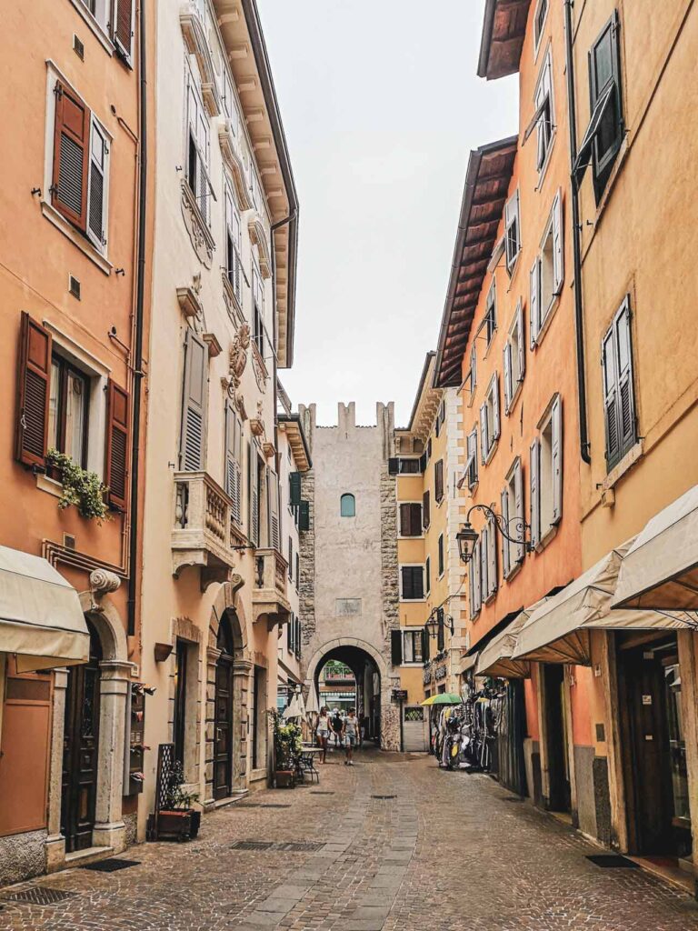 Sehenswürdigkeiten in Riva del Garda am nördlichen Gardasee in Italien: Das Bild zeigt eine Gasse in Riva del Garda mit Blick auf das historische Stadttor Porta San Marco, gesäumt von mediterranen Häusern in Gelb, Terrakotta und Orange