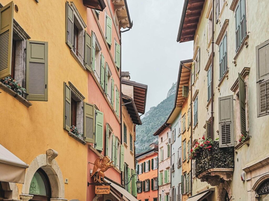 Riva del Garda am nördlichen Gardasee in Italien: Das Bild zeigt die Gasse Via Fiume, gesäumt von typisch italienischen Häusern. Die Häuser sind in mediterranen Farben in Gelb, Terrakotta, Grün und Blau gestrichen und haben hellgrüne Fensterläden.