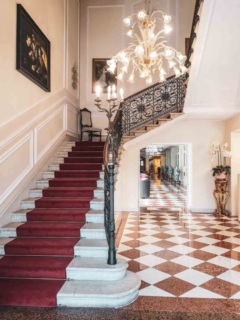 Riva del Garda am nördlichen Gardasee in Italien: Das Bild zeigt die imposante Eingangshalle des Grand Hotel Liberty in mit einem roten Teppich am Treppenaufgang, einem Kronleuchter und Fliesen mit Karomuster in Terrakotta- und Beige-Farben