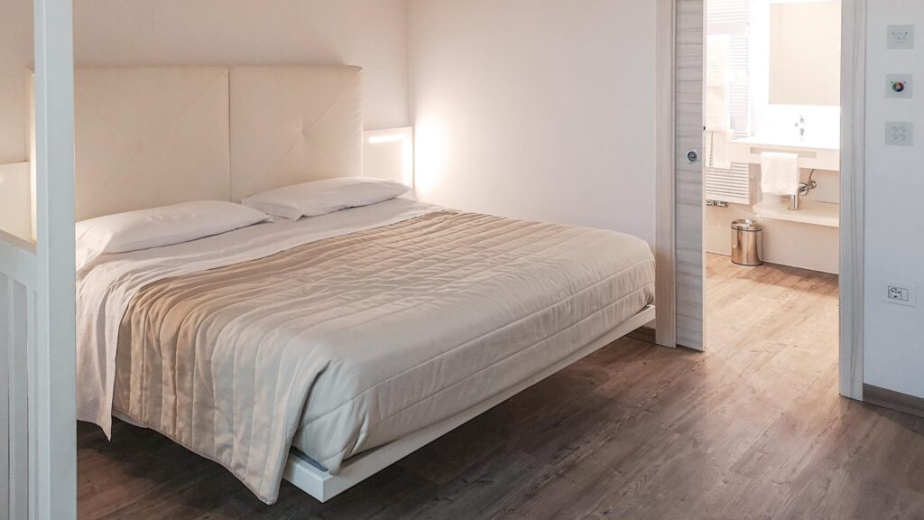 Riva del Garda am nördlichen Gardasee in Italien: Das Bild zeigt eine moderne Suite im Grand Hotel Liberty. Zu sehen ist ein Doppelbett in cremefarben mit dezenter Beleuchtung und hellen Holz- und Weißtönen