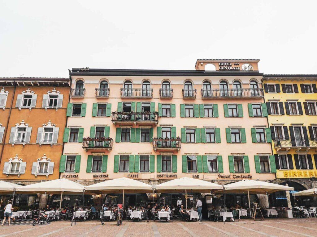 Riva del Garda am nördlichen Gardasee in Italien: Das Bild zeigt die Vorderansicht des Hotel Centrale in der Altstadt von Riva del Garda an der Piazza III Novembre. Das Haus in mediterranem Stil ist cremefarben mit grünen Fensterläden. Im Café sitzen Gäste unter weißen Sonnenschirmen