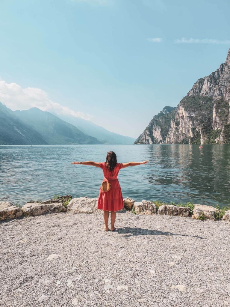 Riva del Garda am nördlichen Gardasee in Italien: Das Bild zeigt Bloggerin Alexandra Baier von Tables & Fables, die mit ausgebreiteten Armen auf einer Aussichtsplattform am Punta Lido in Riva del Garda steht. Sie trägt ein korallfarbenes Sommerkleid und blickt auf den Gardasee und die Berge