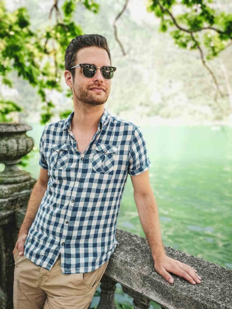 Riva del Garda am nördlichen Gardasee in Italien: Das Bild zeigt Florian Baier von Tables & Fables. Er trägt ein blau-kariertes Hemd und eine Sonnenbrille und steht an der Uferpromenade von Riva del Garda am. Im Hintergrund ist das türkisfarbene Wasser des Gardasees zu sehen