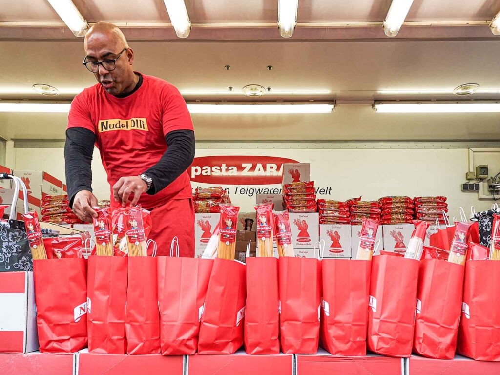 Reisetipps Hamburg: Nudel Olli auf dem Hamburger Fischmarkt verkauft frische Teigwaren am Verkaufsstand mit roten Tüten an der ThekeReisetipps Hamburg: Nudel Olli auf dem Hamburger Fischmarkt verkauft frische Teigwaren am Verkaufsstand mit roten Tüten an der Theke