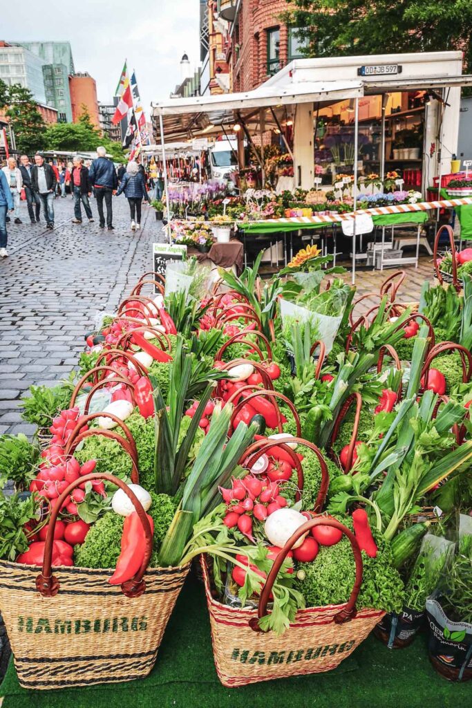 Reisetipps Hamburg: Marktstand auf dem Hamburger Fischmarkt mit bunten Obst- und Gemüsekörben in Weidekörben mit Hamburg-Aufschrift – typische Marktszene und Souvenir- oder Mitbringsel-Idee