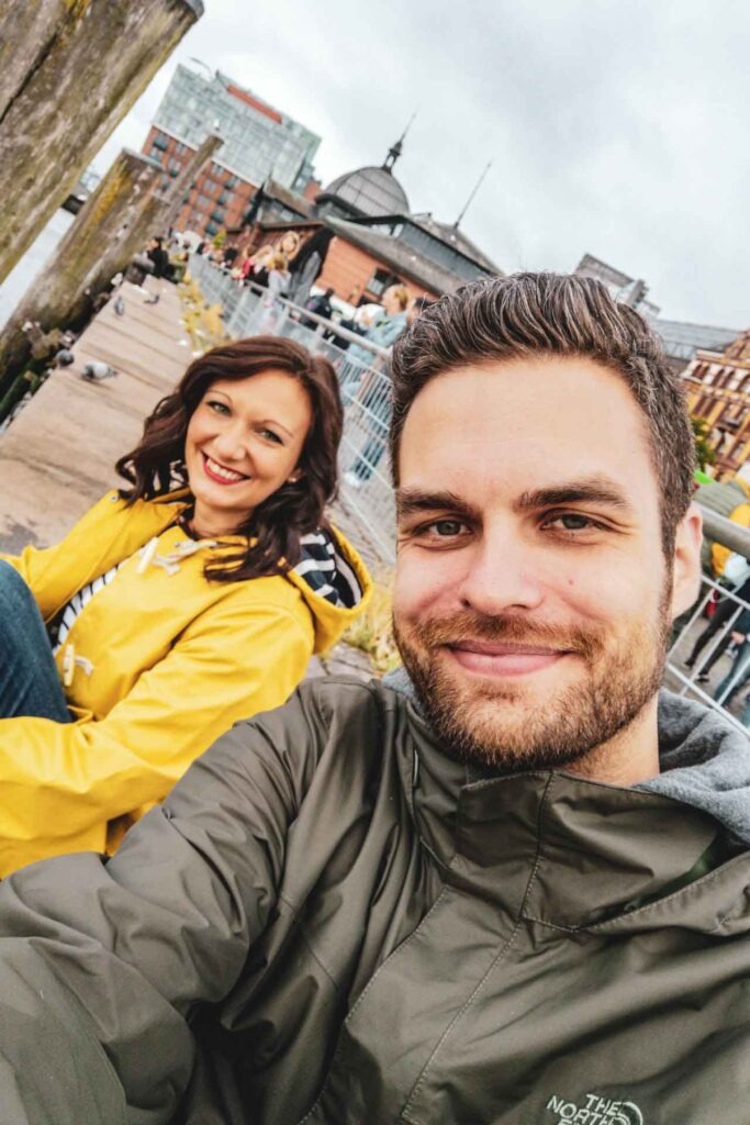 Reisetipps Hamburg: Florian und Alexandra trinken Kaffee an der Uferpromenade des Hamburger Fischmarkts am Elbufer bei grauem Himmel und Regenjacken am Sonntagmorgen