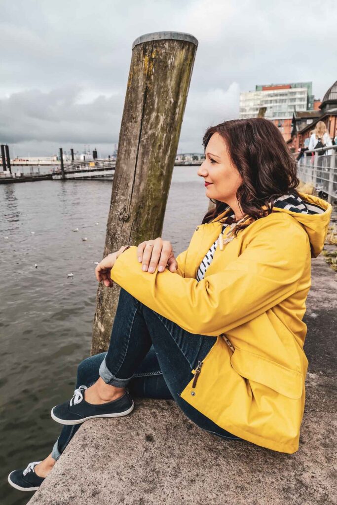 Reisetipps Hamburg: Alexandra in gelber Regenjacke entspannt an der Uferpromenade des Hamburger Fischmarkts am Elbufer bei grauem Himmel am Sonntagmorgen