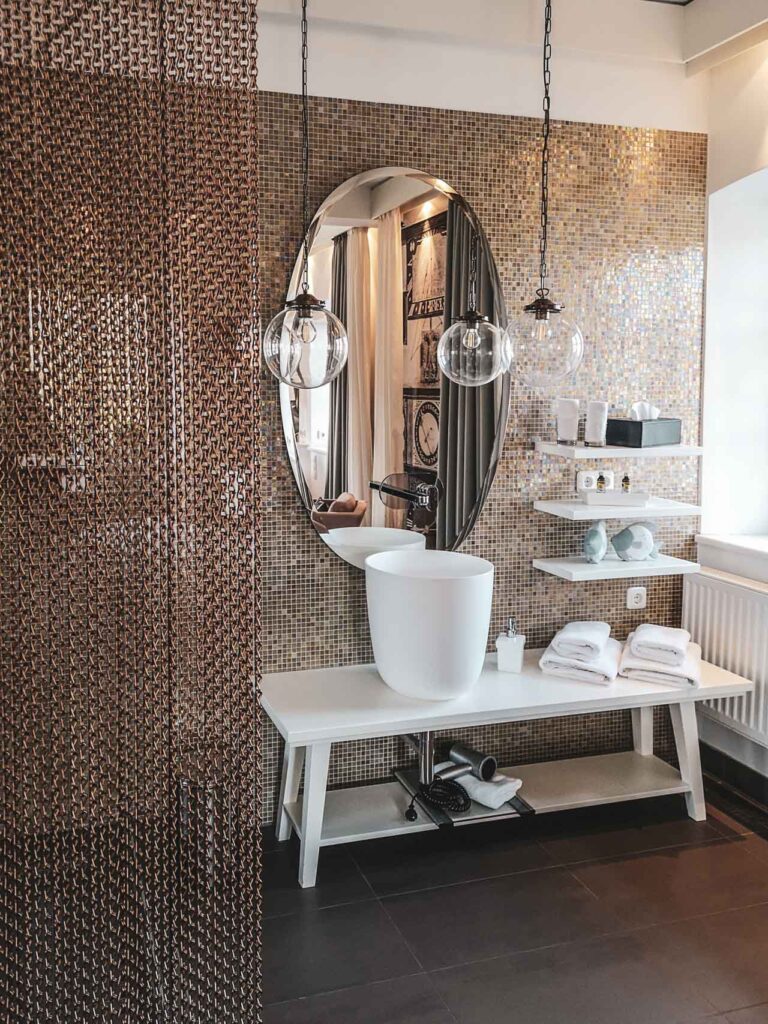 Das offene Badezimmer im Superior Zimmer des Hotels Villa Mittermeier in Rothenburg ob der Tauber in Beige- und Weißtänen. Zu sehen ist ein Spiegel mit Waschtisch in Weiß, davor hängt ein beige-transparenter Vorhang als Raumtrenner