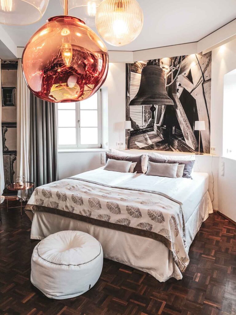 Superior Zimmer im Hotel Villa Mittermeier in Rothenburg ob der Tauber. Das Hotelzimmer ist in Beige-, Rosé und Weißtönen gehalten. Zu sehen sind ein großes Doppelbett mit Tagesdecke und Zierkissen auf der rechten Seite. Im Vordergrund steht ein weißer Fußhocker, eine Lampe im Kupferfarben hängt ins Bild. Am Kopfende hängt eine große Schwarz-Weiß-Fotografie, die eine Kirchglocke umgeben von Fachwerkgauben zeigt