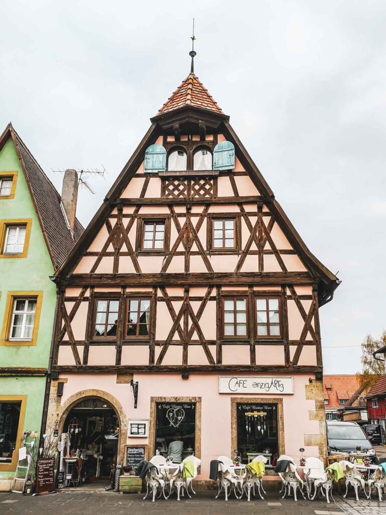 Das gemütliche Café Einzigartig, ein Ladencafé im Vintagestil im historischen Fachwerkhaus in Rosa in Rothenburg ob der Tauber. Davor stehen weiße Gartenmöbel mit bunten Decken