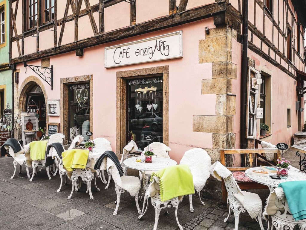 Das Café Einzigartig in Rothenburg ob der Tauber, ein Ladencafé im Vintagestil. Auf dem Bild ist die Fachwerkfassade in Rosa und der Eingang zu sehen, davor stehen Gartenmöbel im Shabby-Chic-Stil in Weiß mit bunten Decken