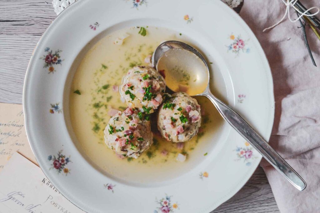Südtiroler Speckknödelsuppe in einem Porzellanteller mit Blumenmuster und Suppenlöffel auf einem rustikalen Holztisch