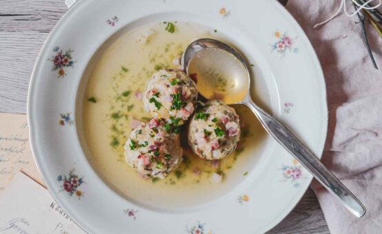 Südtiroler Speckknödelsuppe in einem Porzellanteller mit Blumenmuster und Suppenlöffel auf einem rustikalen Holztisch