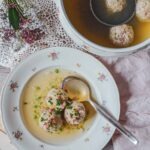 Südtiroler Speckknödelsuppe in einem Porzellanteller mit Blumenmuster und Suppenlöffel auf einem rustikalen Holztisch. Daneben eine traditionelle Suppenschüssel mit Schöpfkelle