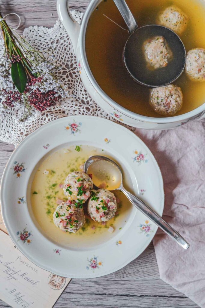 Südtiroler Speckknödelsuppe in einem Porzellanteller mit Blumenmuster und Suppenlöffel auf einem rustikalen Holztisch. Daneben eine traditionelle Suppenschüssel mit Schöpfkelle