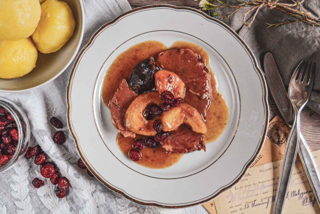 Schlesisches Himmelreich auf einem Porzellanteller mit Goldrand. Daneben stehen eine Schüssel mit schlesischen Klößen, eine Schale mit Cranberrys und Silberbesteck auf einer weißen Spitzendecke