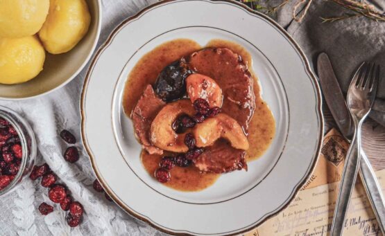Schlesisches Himmelreich auf einem Porzellanteller mit Goldrand. Daneben stehen eine Schüssel mit schlesischen Klößen, eine Schale mit Cranberrys und Silberbesteck auf einer weißen Spitzendecke
