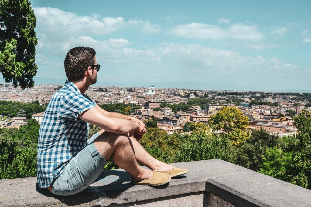 Italien im Sommer: Der Aussichtspunkt Terrazza Belvedere Aventino auf dem Aventin, dem südlichsten der sieben Hügel in Rom. Florian von Tables & Fables sitzt auf einer grauen Steinmauer und blickt auf die Stadt