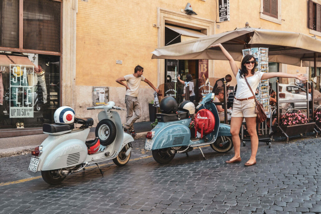 Italien im Sommer: Alexandra von Tables & Fables steht vor zwei typisch italienischen Vespas in Grau und Hellblau vor einer Bar in Rom