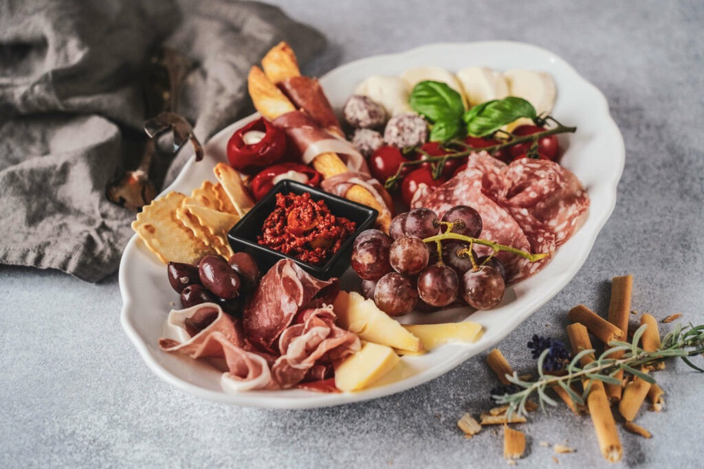 Antipastiplatte mit typisch italienischen Produkten: Mozzarella, Salami, Prosciutto, Parmaschinken, Weintrauben, Grissini und Tomaten auf einer Servierplatte aus Porzellan. Im Hintergrund ist eine graue Stoffserviette zu sehen