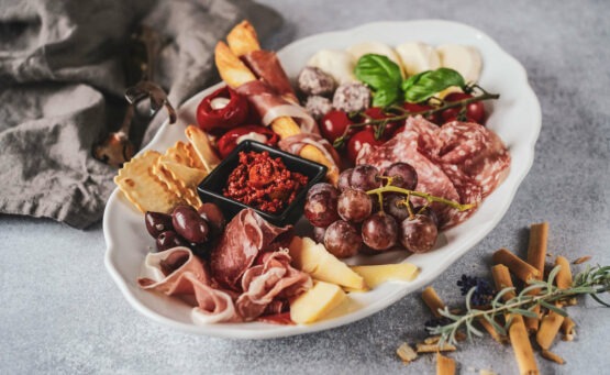 Antipastiplatte mit typisch italienischen Produkten: Mozzarella, Salami, Prosciutto, Parmaschinken, Weintrauben, Grissini und Tomaten auf einer Servierplatte aus Porzellan. Im Hintergrund ist eine graue Stoffserviette zu sehen