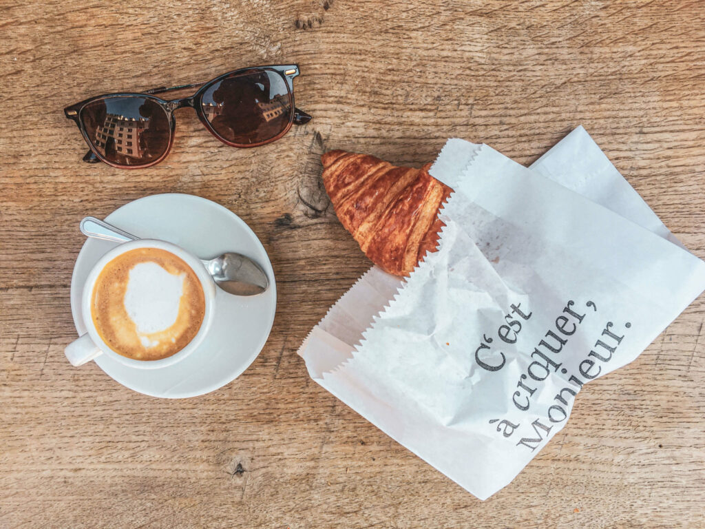 Italienfeeling für zu Hause: Typische italienisches Frühstück mit Cappuccino und Cornetto auf einem Holztisch, daneben liegt eine Sonnenbrille
