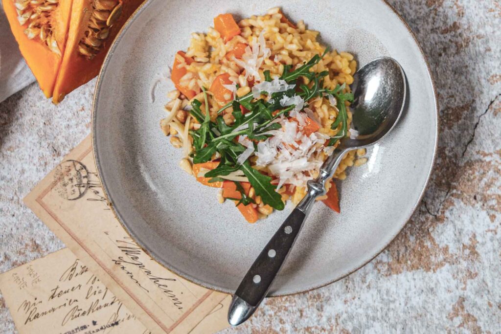 Ein geschmiedeter Esslöffel mit schwarzem Griff und Nieten. Er liegt in einem tiefen sandfarbenen Steingutteller mit Kürbisrisotto auf einem Steinuntergrund mit Vintage-Postkarten
