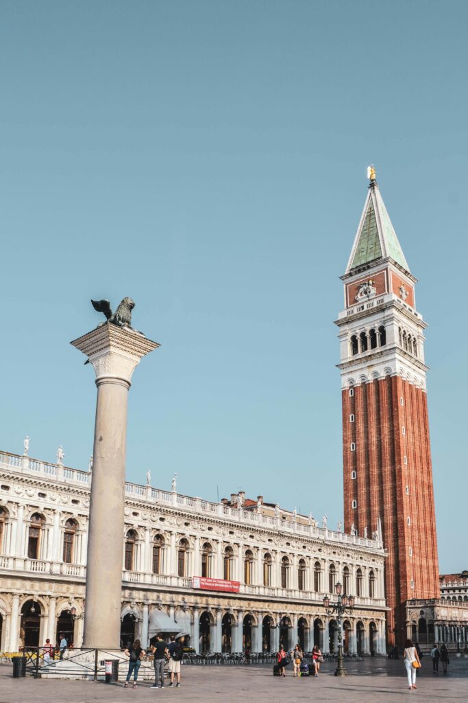 Die Piazza San Marco in Venedig am Morgen, getaucht in warmes Sonnenlicht, mit Blick auf den Markusturm sowie die Löwenstatue Colonna di San Marco.