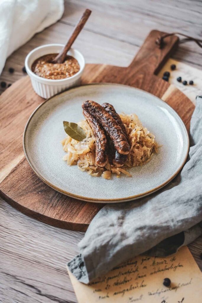 Drei Nürnberger Rostbratwürste auf Sauerkraut, serviert auf einem Steingutteller auf einem rustikalen Holzbrett. Daneben stehen eine kleine Schüssel mit süßem Weißwurstsenf und ein Holzlöffel, daneben liegen eine graue Serviette und Vintage-Postkarten.