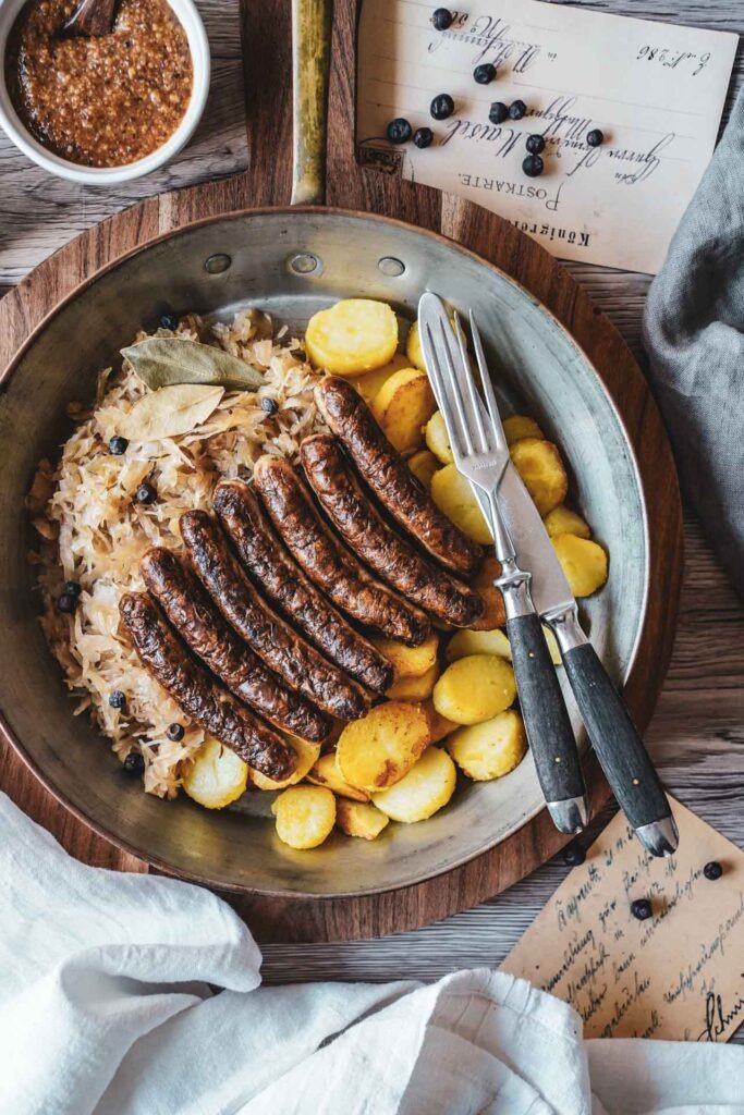Nürnberger Rostbratwürste mit Bratkartoffeln, Sauerkraut und süßem Weißwurstsenf, serviert in einer Kupferpfanne auf einem rustikalen Holzbrett. Daneben liegen Besteck, eine weiße Tischdecke, eine Vintage-Postkarte und Wacholderbeeren.