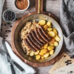 Nürnberger Rostbratwürste mit Bratkartoffeln, Sauerkraut und süßem Weißwurstsenf, serviert in einer Kupferpfanne auf einem rustikalen Holzbrett. Daneben liegen Besteck, eine weiße Tischdecke, eine Vintage-Postkarte und Wacholderbeeren.