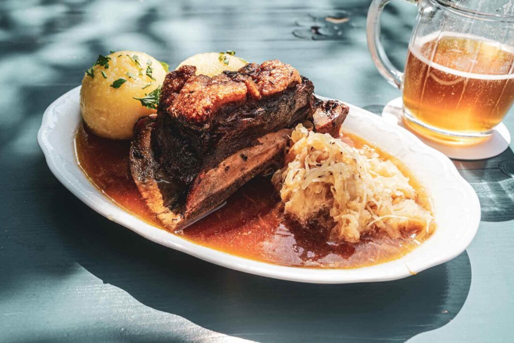 Foodtipps für Bayreuth: Der Biergarten auf der Thema mit regionalen Spezialitäten. Das Bild zeigt einen ovalen Teller mit traditionellem Krustenschäufele, zwei Klößen, Sauerkraut und Bratensauce auf einem blau gestrichenen Tisch, im Hintergrund ein Glaskrug mit Bernsteinfarbenem Bier