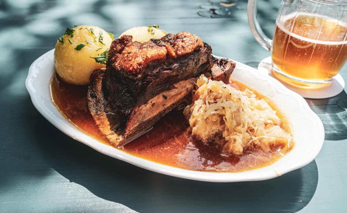 Foodtipps für Bayreuth: Der Biergarten auf der Thema mit regionalen Spezialitäten. Das Bild zeigt einen ovalen Teller mit traditionellem Krustenschäufele, zwei Klößen, Sauerkraut und Bratensauce auf einem blau gestrichenen Tisch, im Hintergrund ein Glaskrug mit Bernsteinfarbenem Bier