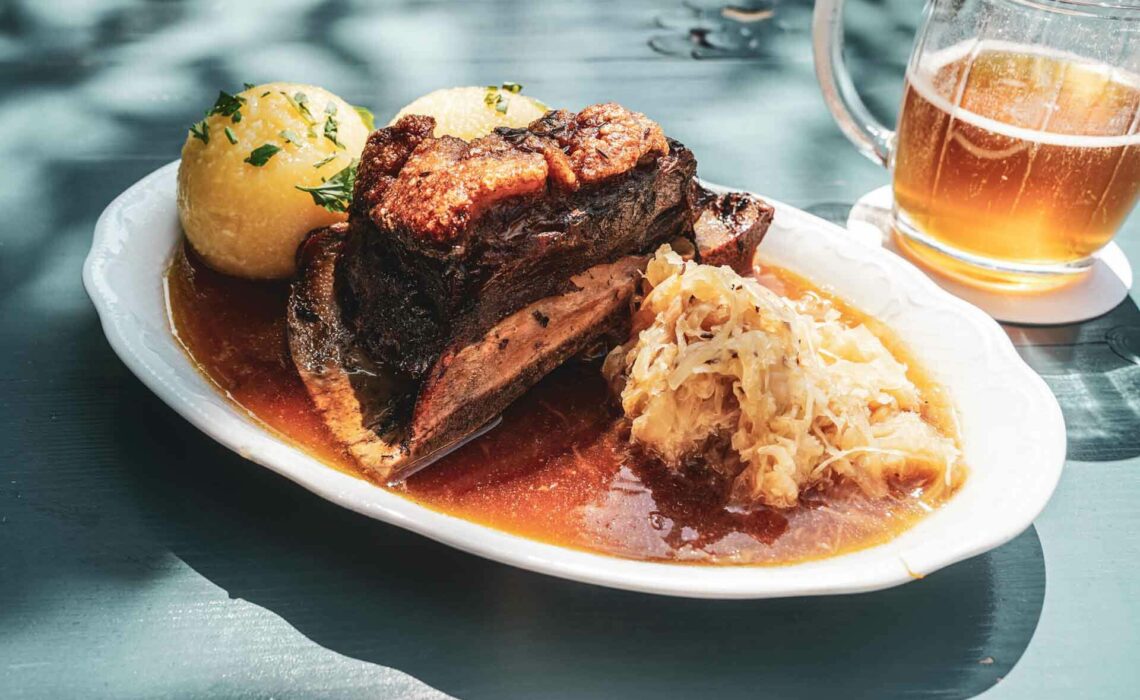 Restauranttipps für Bayreuth: Der Biergarten auf der Thema mit regionalen Spezialitäten. Das Bild zeigt einen ovalen Teller mit traditionellem Krustenschäufele, zwei Klößen, Sauerkraut und Bratensauce auf einem blau gestrichenen Tisch, im Hintergrund ein Glaskrug mit Bernsteinfarbenem Bier