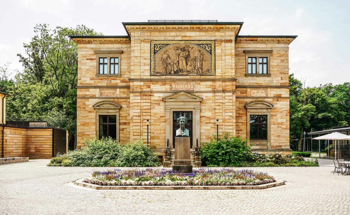 Sehenswürdigkeiten in Bayreuth: Die Vorderseite von Haus Wahnfried, das als ehemaliges Wohnhaus Richard Wagners Teil des Richard Wagner Museums ist. Vor der Sandsteinvilla ist die Büste König Ludwigs II. in einem Blumenbeet zu sehen, rechts und links weitere Museumsgebäude, im Hintergrund sind grüne Bäume zu sehen