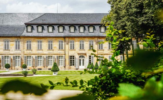 Sehenswürdigkeiten in Bayreuth: Das Neue Schloss im Sommer. Blick vom Hofgarten durch grüne Blätter hindurch auf das barocke Sandsteingebäude mit himmelblauen Fensterläden und weißen Sprossenfenstern