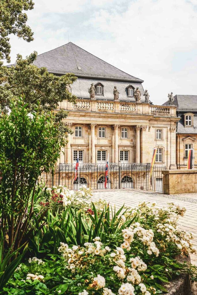 Sehenswürdigkeiten in Bayreuth: Das Markgräfliche Opernhaus. Blick von der Aussichtsplattform des Mariengärtleins auf das Sandsteingebäude, vor dem verschiedene Länderflaggen aufgestellt sind. Im Vordergrund sind Blumenbeete mit weißen Rosen zu sehen