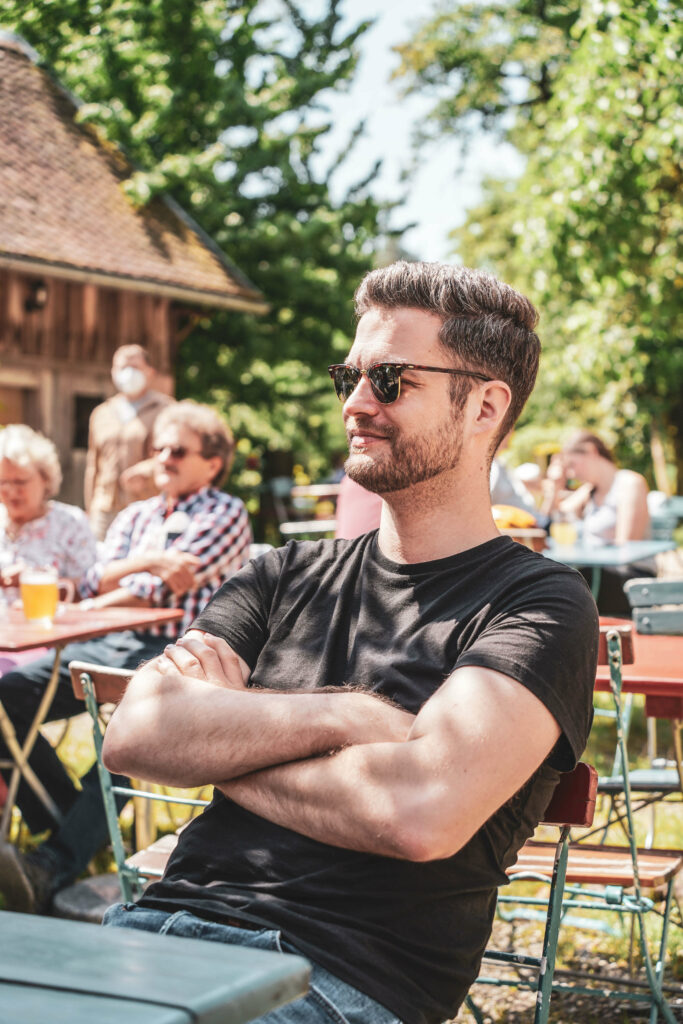 Flo von Tables & Fables sitzt im Biergarten der Theta in Bindlach bei Bayreuth an einem schattigen Platz auf einer blau gestrichenen Bierbank. Er traegt ein schwarzes T-Shirt, eine Sonnenbrille, hat die Arme vor der Brust verschraenkt und blickt laechelnd zur Seite. Im Hintergrund sind eine Scheune, hochgewachsene Baeume und weitere Bierbaenke mit Besuchern zu sehen