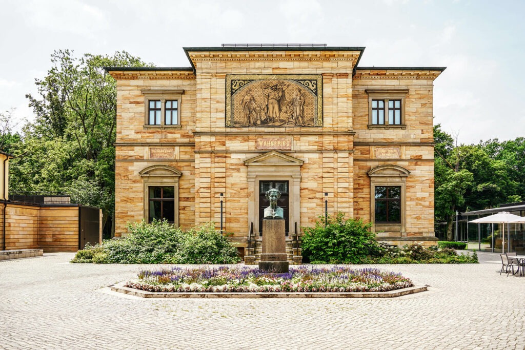 Haus Wahnfried, das ehemalige Wohnhaus des Komponisten Richard Wagner in Bayreuth am Hofgarten, beherbergt heute das Richard Wagner Museum. Das Bild wurde um Sommer aufgenommen und zeigt das Haus mit grünen Bäumen um Hintergrund sowie Blumen im davor liegenden Beet, das eine Büste Ludwigs des II. ziert.