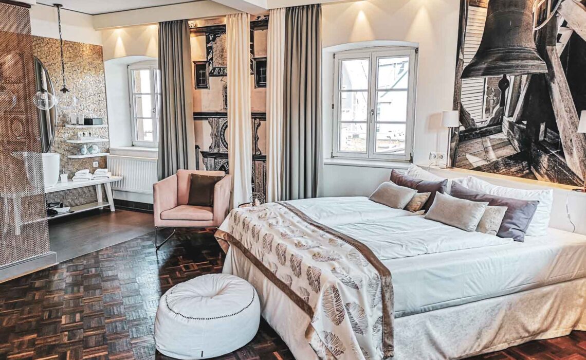 Superior Zimmer im Hotel Villa Mittermeier in Rothenburg ob der Tauber. Das Hotelzimmer ist in Beige-, Rosé und Weißtönen gehalten. Zu sehen sind ein großes Doppelbett mit Tagesdecke und Zierkissen auf der rechten Seite. Im Vordergrund steht ein weißer Fußhocker, im Hintergrund sind ein rosafarbener Sessel sowie ein Teil des offenen Badezimmers zu sehen