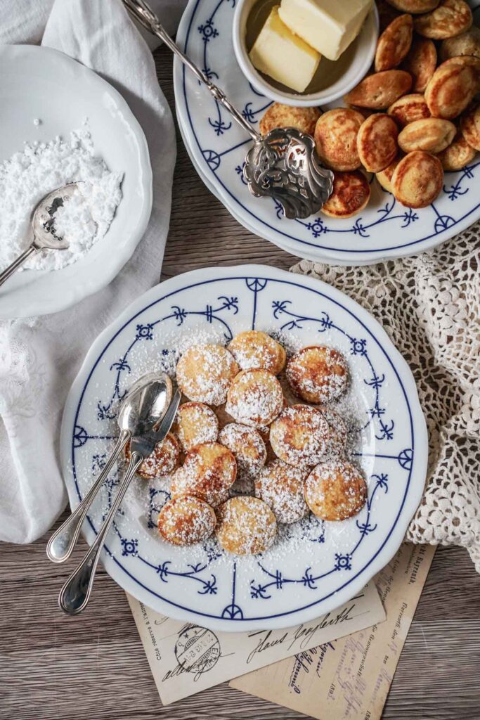 Holländische Poffertjes mit Puderzucker und Butter auf einem Porzellanteller mit Friesenmuster, serviert auf einem rustikalen Untergrund mit heller Häkeldecke
