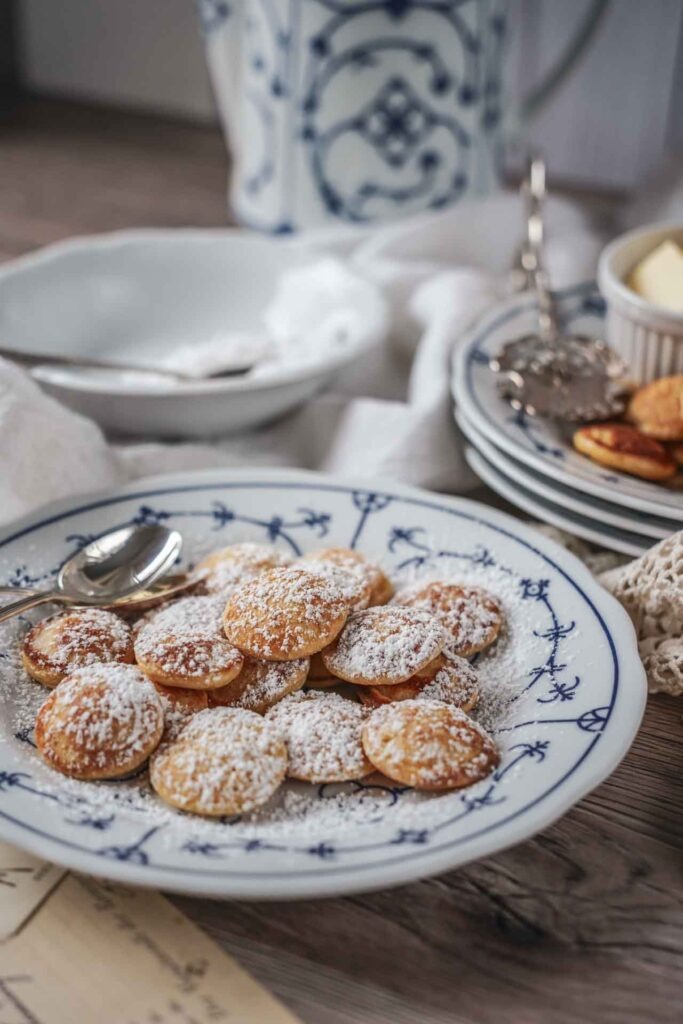 Holländische Poffertjes mit Puderzucker und Butter auf einem Porzellanteller, serviert auf einem rustikalen Untergrund mit heller Decke. Im Hintergrund steht eine Teekanne.