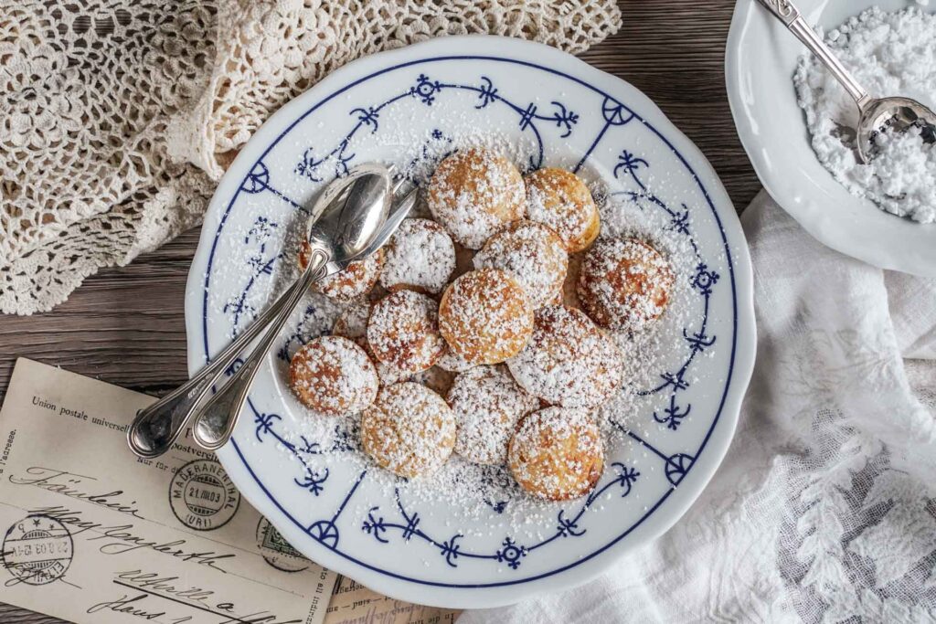 Holländische Poffertjes mit Puderzucker auf einem Porzellanteller mit Friesenmuster, serviert auf einem rustikalen Untergrund mit heller Decke