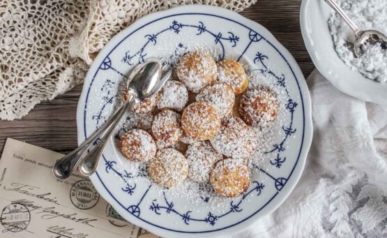 Holländische Poffertjes mit Puderzucker auf einem Porzellanteller mit Friesenmuster, serviert auf einem rustikalen Untergrund mit heller Decke