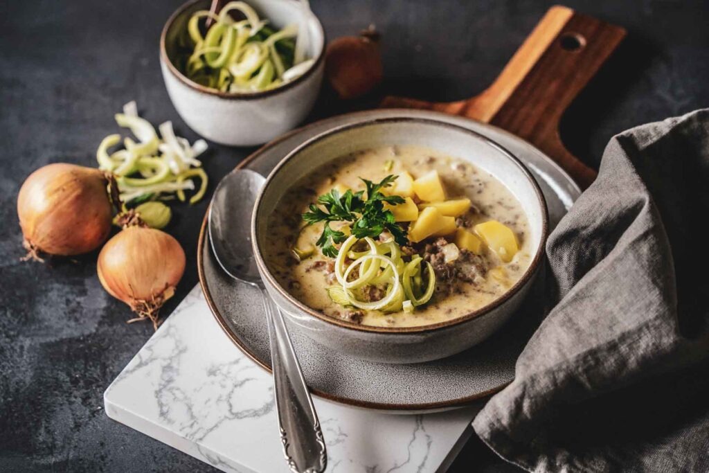 Käse-Lauch-Suppe in einer Steingutschüssel auf einem Servierbrett. Im Vordergrund liegt eine graue Stoffserviette, im Hintergrund steht eine kleine Schüssel mit geschnittenen Lauchringen