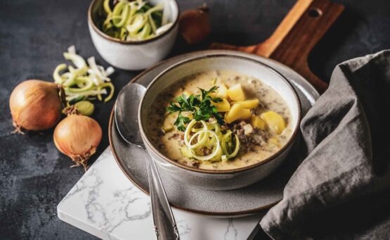 Käse-Lauch-Suppe in einer Steingutschüssel auf einem Servierbrett. Im Vordergrund liegt eine graue Stoffserviette, im Hintergrund steht eine kleine Schüssel mit geschnittenen Lauchringen