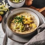 Käse-Lauch-Suppe in einer Steingutschüssel auf einem Servierbrett. Im Vordergrund liegt eine graue Stoffserviette, im Hintergrund steht eine kleine Schüssel mit geschnittenen Lauchringen