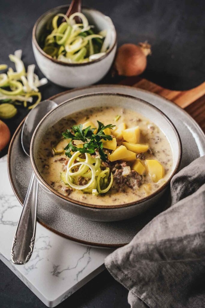 Käse-Lauch-Suppe in einer Steingutschüssel auf einem Servierbrett. Im Vordergrund liegt eine graue Stoffserviette, im Hintergrund steht eine kleine Schüssel mit geschnittenen Lauchringen